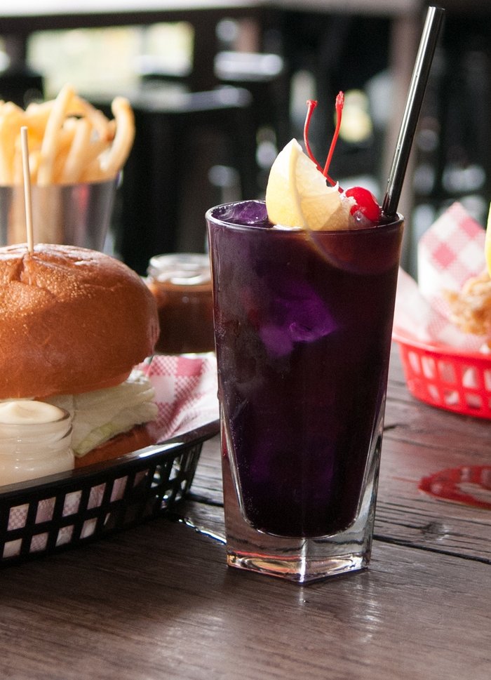 A tall purple cocktail with a lemon slice and cherries beside a burger and fries